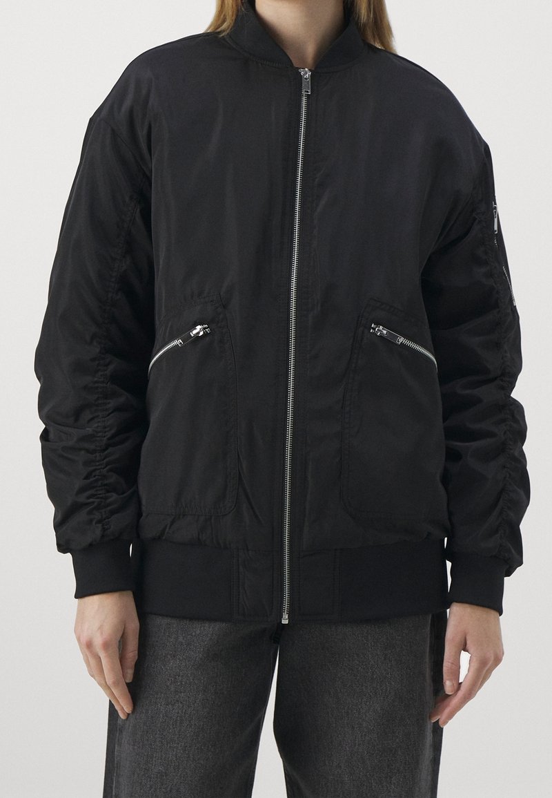 Personne portant une veste bomber noire avec des poches zippées et une fermeture éclair à l'avant, associée à un pantalon gris foncé, sur un fond uni.