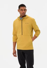 Gelb-geschlechtete Kapuzensweatshirt mit einem Halb-Zip-Design, schwarzem Reißverschluss, strukturiertem Stoff und Fronttasche. Kombiniert mit schwarzen Sporthosen.