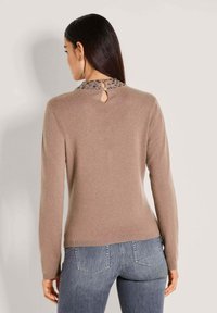 Vrouw met lang, donker haar draagt een beige trui met versierde kraag en een sleutelgatknopsluiting, in combinatie met verouderde blauwe jeans, en kijkt weg.