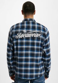 Kék és fekete kockás flanel ing, hosszú ujjakkal, lekerekített hemgel, és a háton hímzett "Rocawear" felirattal.