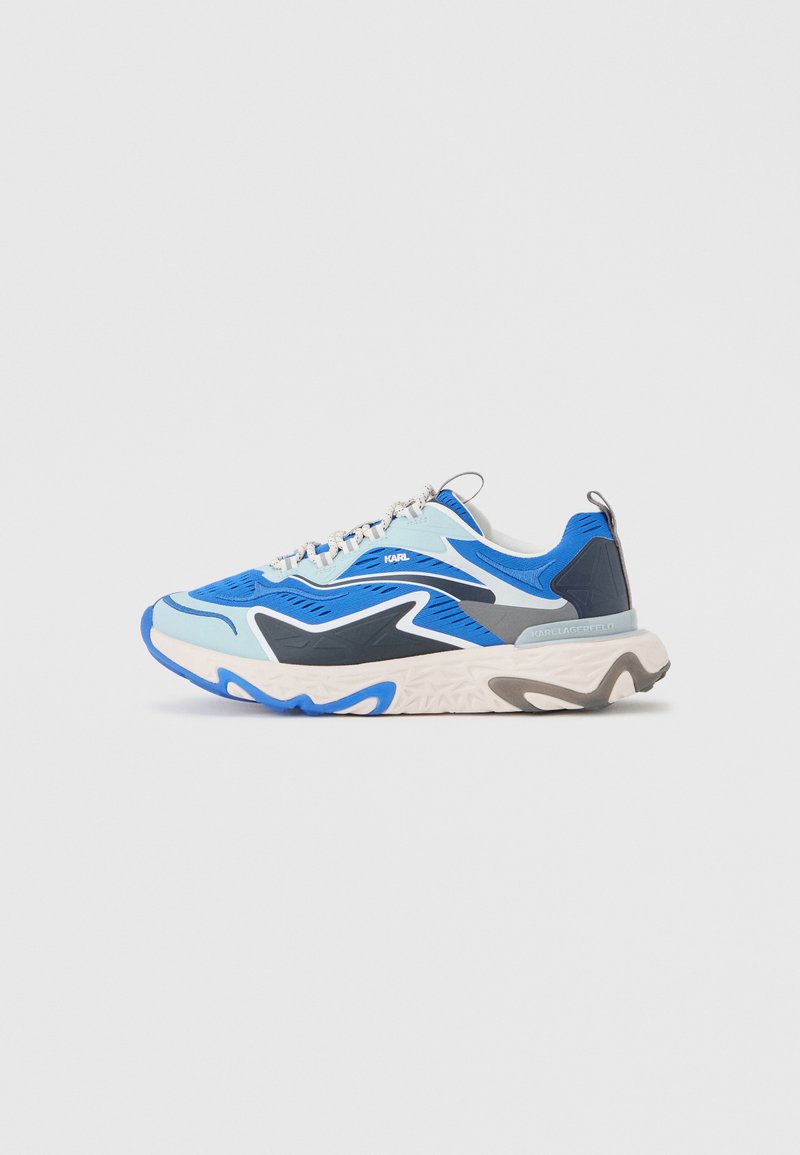 Zapatilla deportiva azul, negra y blanca con patrones ondulados, suela gruesa y tira para tirar en el talón, mostrada sobre fondo gris liso.