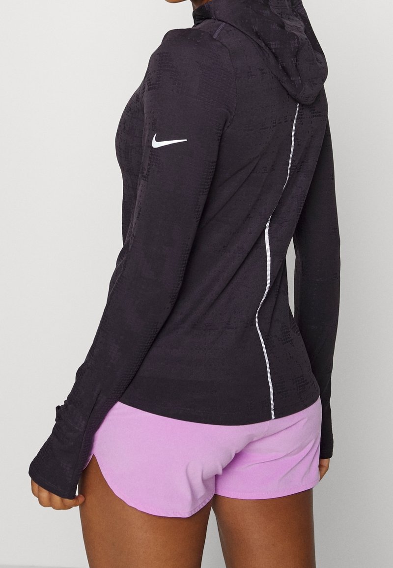 Personne portant un sweat à capuche noir à manches longues avec des trous pour les pouces et un short violet clair, vue de dos sur un fond uni.
