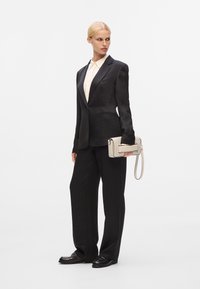 Blazer nero su misura in tessuto testurizzato, abbinato a pantaloni neri a gamba larga. Il modello tiene una borsa chiara con dettagli perforati.