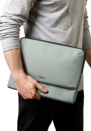 Housse d'ordinateur portable gris clair en tissu texturé, avec un design plat, une fermeture éclair noire et un logo discret, tenue dans une main.