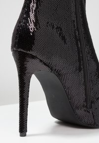 Černá vysoká kozačka na podpatku s texturou z flitrů, zužujícím se tvarem a zipem na boku. Úzký jehlový podpatek dodává na eleganci.
