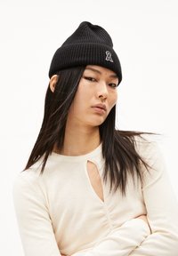 Zwarte geribbelde gebreide beanie met een klein wit logo. Draagbaar door een model met steil zwart haar, gecombineerd met een crèmekleurige top met een uitsnede.