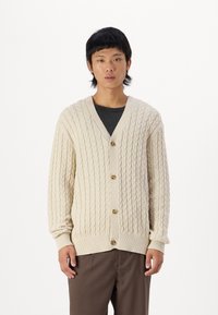 Strikket kremfarget cardigan med teksturert kabeldesign, V-hals, fem-knappers lukking, ribbestrikkede ermer og kant, båret over en mørk topp og brune bukser.