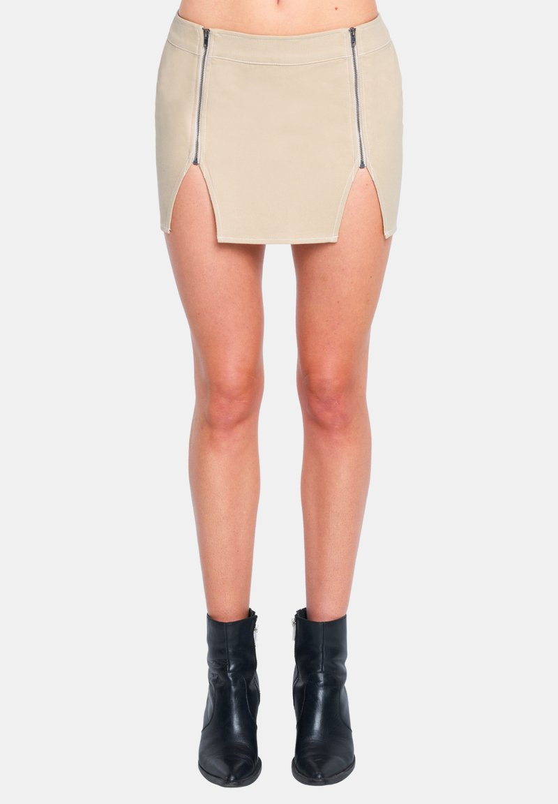 Beige Minirock mit zwei vorderen Reißverschlüssen, seitlichen Schlitzen und sichtbaren Nähten. Kombiniert mit schwarzen Knöchelstiefeln.