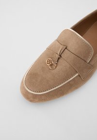 Brown suede loafers featuring a bow detail and a gold accent. Rounded toe with contrasting stitching and a textured surface.