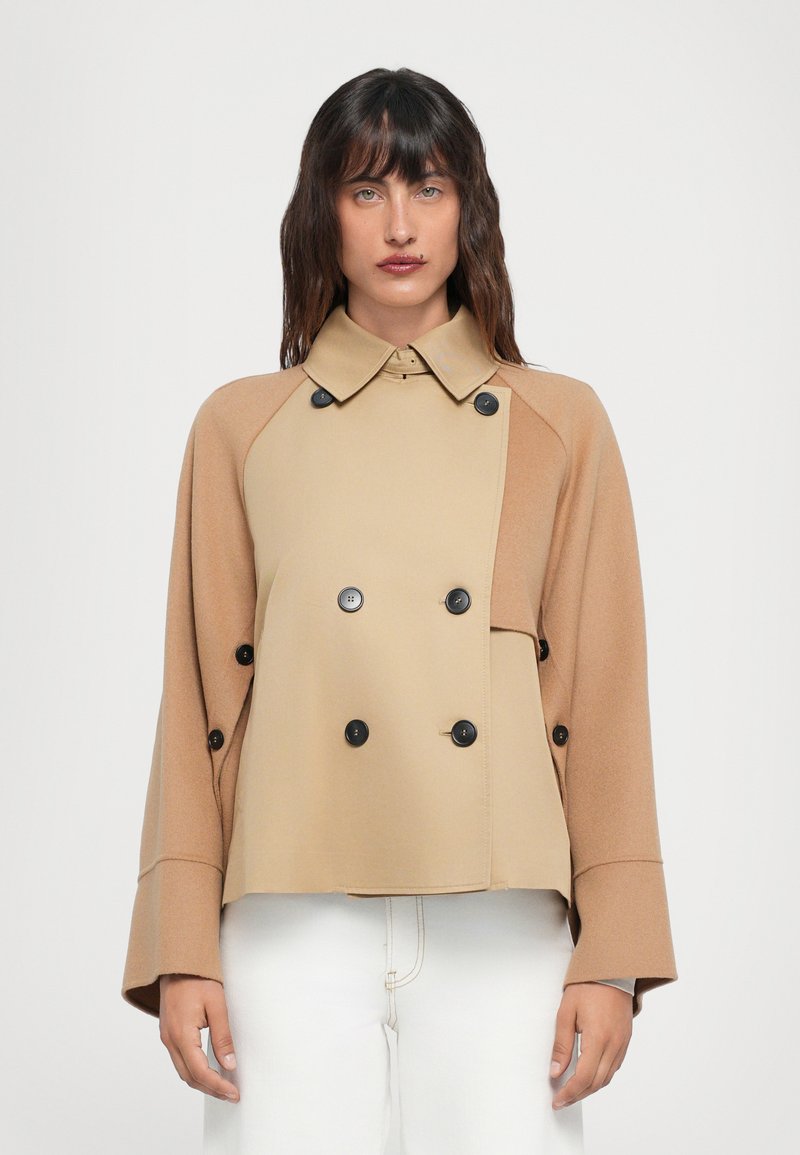 Double-breasted jacket in beige and tan fabric, featuring a sharp collar, large black buttons, and bell sleeves. Textured, contemporary design.