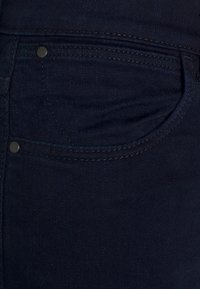 Tela de mezclilla azul oscura con un bolsillo frontal visible. Presenta un botón de metal y un fino cosido a lo largo de los bordes. Textura suave.
