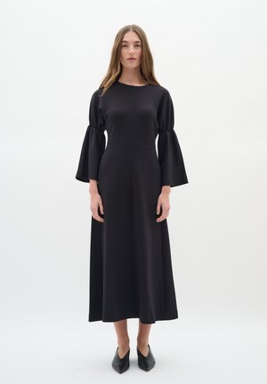 Schwarzes Langarmkleid mit ausgestellten Ärmeln, aus glattem Stoff gefertigt. Verfügt über ein tailliertes Oberteil und einen fließenden Rock, ergänzt durch schwarze spitze Schuhe.