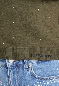Maglione lavorato a maglia verde oliva con piccole decorazioni in oro rosa e orlo a costine, etichettato "Pepe Jeans" in blu scuro. Abbinato a pantaloni corti in denim.