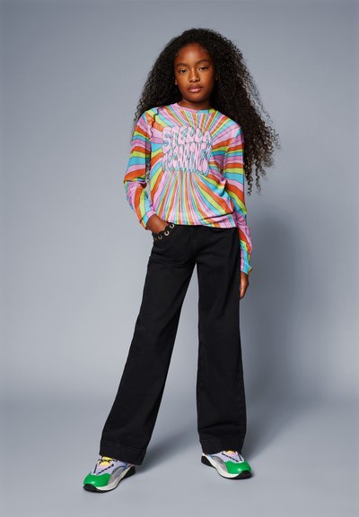 Camicia a maniche lunghe a righe multicolori con testo grafico, pantaloni neri a gamba dritta e scarpe da ginnastica multicolori con dettagli verdi.
