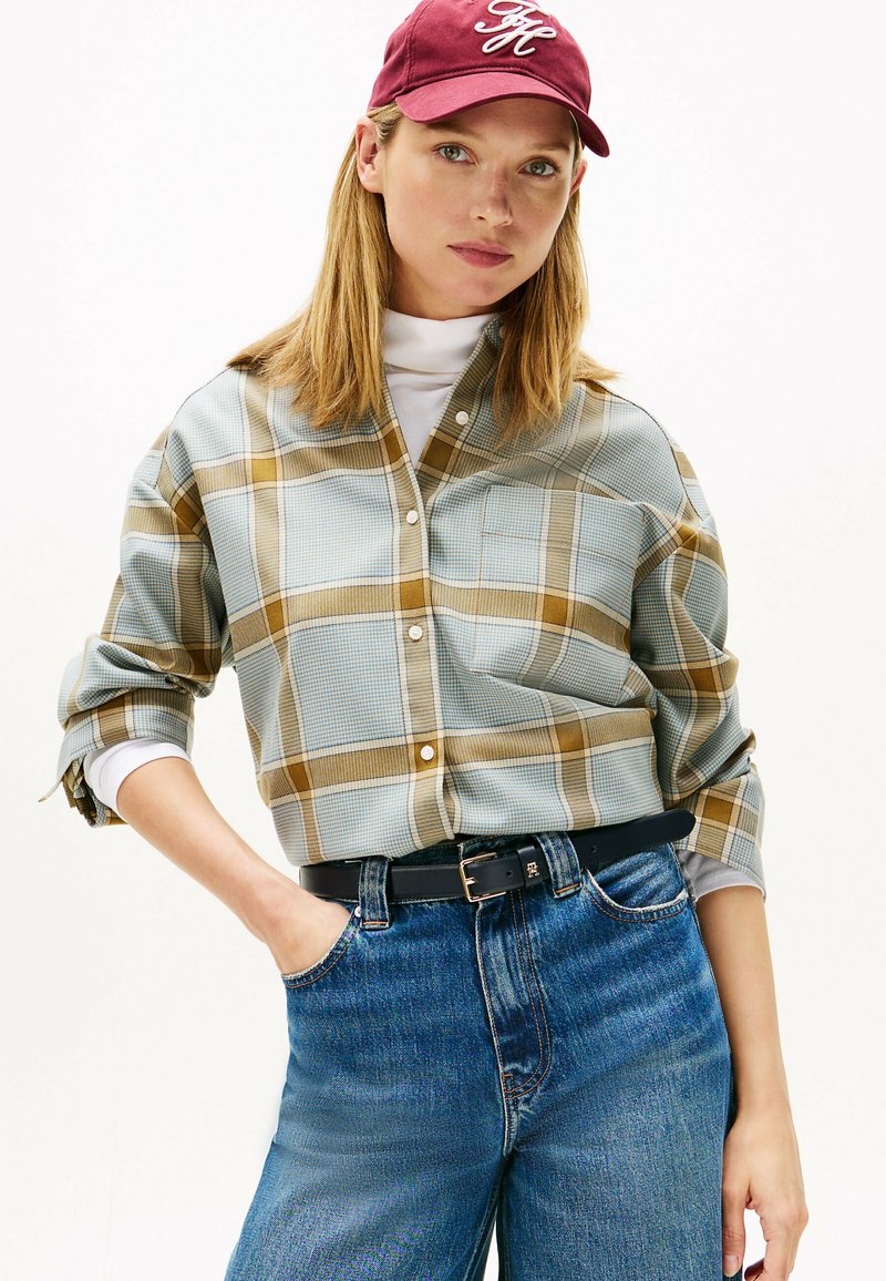 Chemise à boutons avec un motif à carreaux bleu et marron, manches retroussées et une poche poitrine. Portée avec un jean bleu taille haute et une casquette bordeaux.