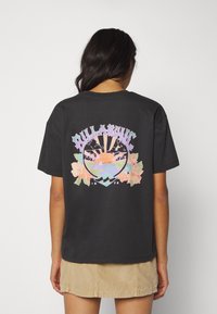 Black cotton t-shirt featuring a colorful graphic on the back, depicting a beach scene with palms and flowers, with "BILLABONG" text above.