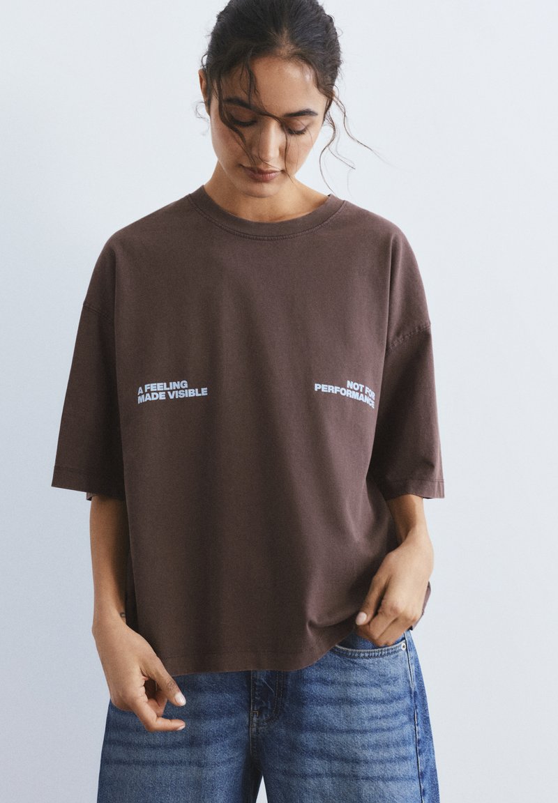 Fiatal nő laza sötét hajjal, túlméretezett barna pólót fehér felirattal és kék farmert visel, lefelé néz egy egyszerű háttér előtt.