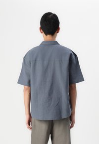 Chemise à manches courtes en tissu gris foncé, dotée d'un col classique et d'un ourlet arrondi, associée à un pantalon de couleur claire.
