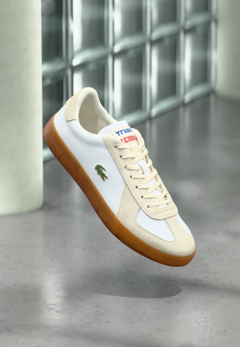 White leather sneaker with beige suede accents, creamy laces, and a brown rubber sole. Features a green logo on the side and colorful branding.