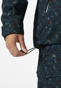 Kurtka wodoodporna z niebieskim abstrakcyjnym wzorem, z regulowanym ściągaczem u dołu i zamkniętą na zamek kieszenią z przodu. Teksturowana powierzchnia materiału.