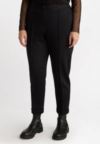 Pantalons noirs ajustés avec une texture lisse, dotés d'une fermeture à bouton à l'avant, de coutures visibles et de ourlets roulés, associés à des bottes noires.