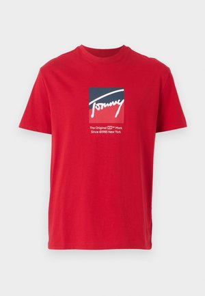 Camiseta de algodón roja con un gráfico cuadrado que presenta "Tommy" en cursiva blanca, rodeado de acentos en azul marino y rojo, y etiquetada con "La Marca Original."