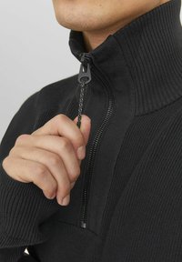 Czarny sweter z zamkiem na szyi o prążkowanej fakturze. Posiada wysoki kołnierz i zamek z ozdobnym pullerem. Zbliżenie na rękę trzymającą zamek.