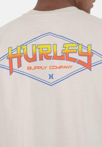 Camiseta de color beige claro con un gráfico colorido en la parte de atrás, con "HURLEY" en un texto negrita amarillo y naranja dentro de un contorno de rombo azul.