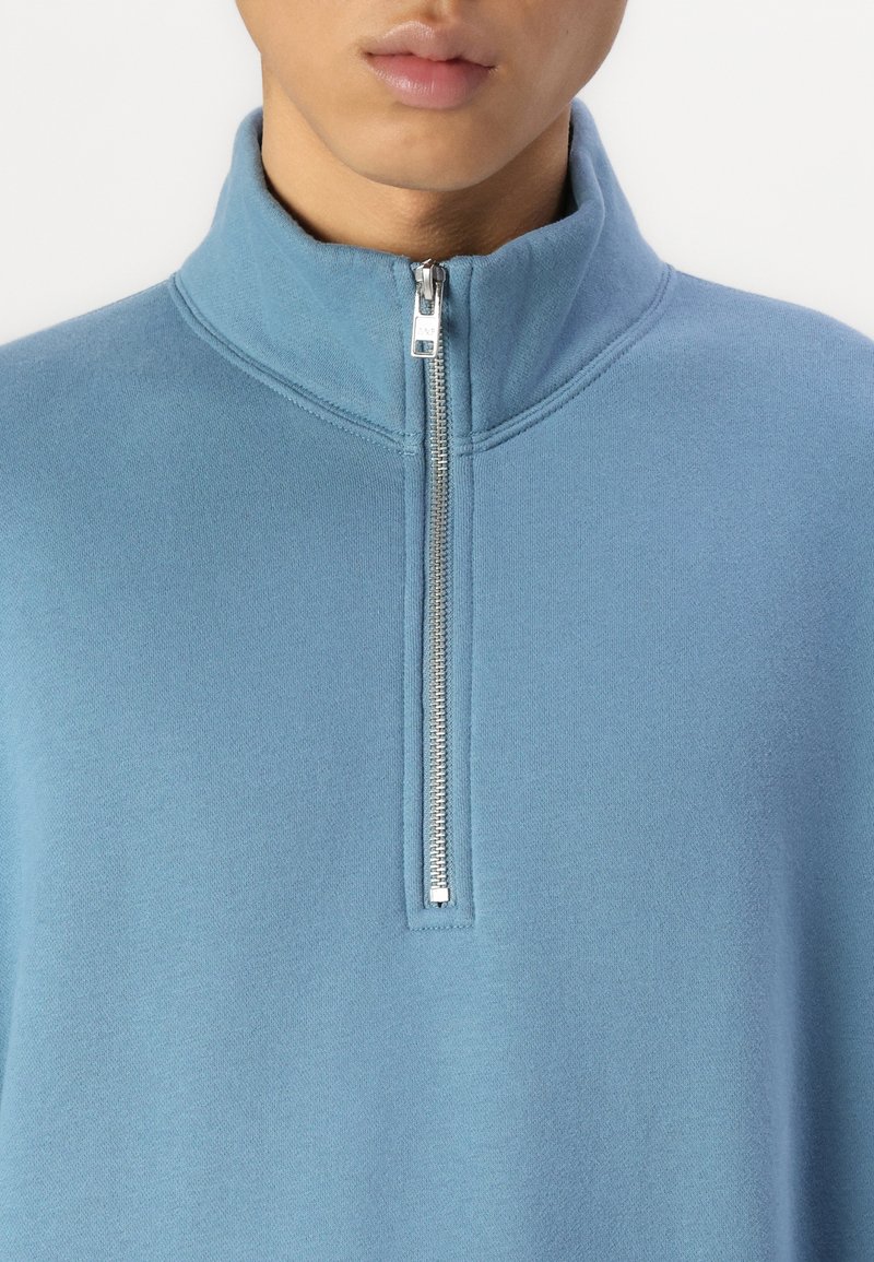 Lichtblauwe ritspullover met hoge kraag, voorzien van een zilveren rits en een zachte, gestructureerde stof. Minimalistisch ontwerp met een ontspannen pasvorm.