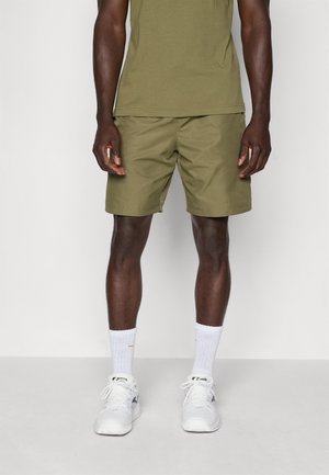 Pantaloni corti color khaki con una texture liscia e una vestibilità rilassata, abbinati a scarpe da ginnastica bianche e calzini, creando un look casual sportivo.