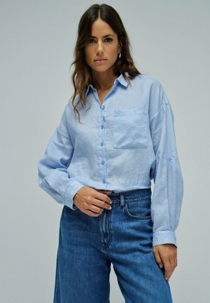 Mujer con cabello largo y marrón, vestida con una camisa azul claro abotonada y jeans azules de cintura alta, de pie contra un fondo liso.