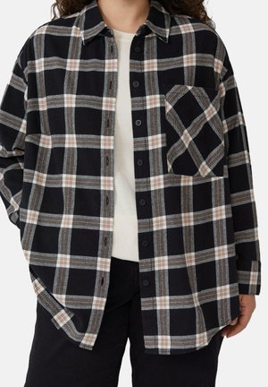 Person iført en sort, grå og beige ternet skjorte med knapper over en hvid top og sorte bukser, stående med den ene hånd i lommen.