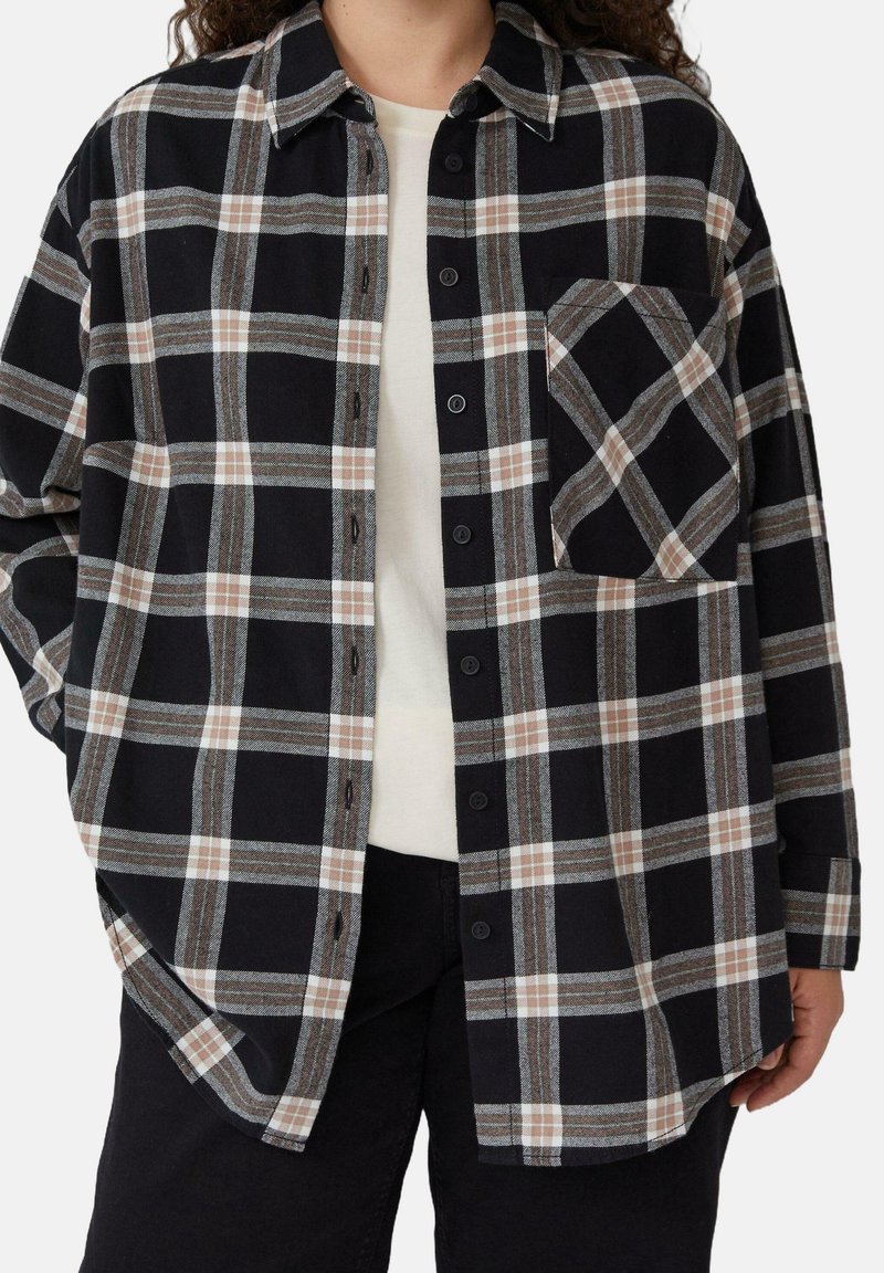 Personne portant une chemise à carreaux noire, grise et beige boutonnée sur un haut blanc et un pantalon noir, debout avec une main dans la poche.