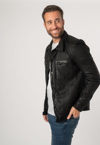 Giacca di pelle nera con chiusura a bottoni, taschino sul petto singolo e dettagli a coste sulle spalle, indossata sopra una camicia bianca e jeans blu.