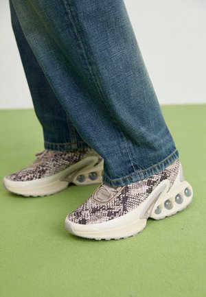 Stopy w sneakersach w wzór skóry węża z przezroczystymi, amortyzującymi powietrzem podeszwami, zestawione z niebieskimi dżinsami na zielonej podłodze.