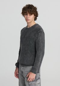 Maglione grigio a coste con maniche lunghe e scollatura a girocollo. Il maglione presenta una trama sottile e dettagli a strisce verticali in contrasto.