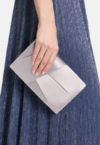 Lichtgrijze satijnen clutch met geplooid ontwerp, vastgehouden in een hand, tegen een structuurrijke donkergroene jurk met metalen draden.
