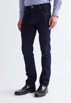 Hombre vestido con vaqueros ajustados azul oscuro, camisa azul clara abotonada y mocasines de cuero negros, de pie frente a un fondo liso.