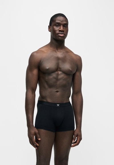 Homme musclé debout portant un boxer noir sur un fond clair uni, regardant devant avec une expression neutre.