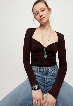 Femme portant un haut noir à manches longues froncé, un jean bleu et des bijoux assortis en pierres bleues sur le collier, le bracelet et la bague.