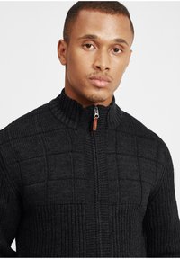 Maglione nero con zip, caratterizzato da un design a trama a griglia. Collo alto, dettagli a coste e zip liscia.
