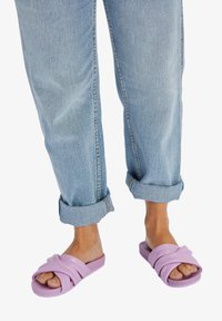 Lavendelkleurige verstelbare sandalen met brede, overlappende banden, gedragen bij lichtblauwe jeans met opgerolde manchetten, die de zachte textuur en het ontspannen ontwerp benadrukken.