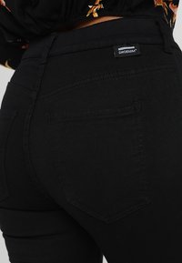 Jeans de mezclilla negra con un diseño ajustado, que presentan dos bolsillos traseros y una etiqueta con la marca "DRDENIM" en la cintura. Textura suave.