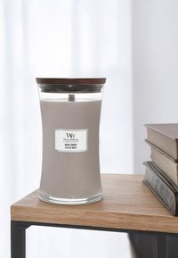 Šedá svíčka v průhledném skleněném držáku se dřevěným víkem. Na etiketě je nápis "Dřevěný kouř." Dřevěný knot je viditelný uvnitř; položeno na světle dřevěném stole.