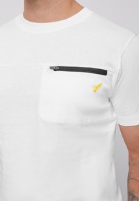 White short-sleeve t-shirt with a black zippered chest pocket and yellow embroidered bird logo. Smooth cotton fabric, round neckline.