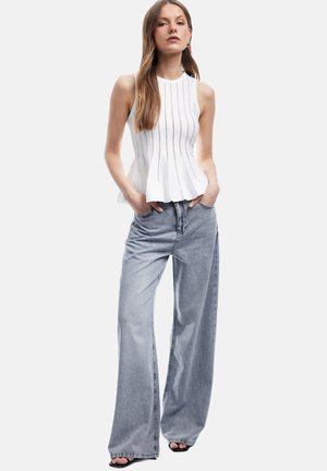 Weißes, ärmelloses Top mit vertikalen Rippstrukturen und einem ausgestellten Saum, kombiniert mit weit geschnittenen, hellgrauen Jeans und schwarzen, offenen High Heels.