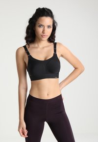 Svart sports-bh med brede stropper og et teksturert design. Har et støttende bånd og justerbare detaljer. Brukt med mørke leggings.