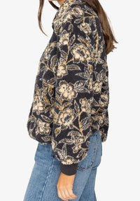 Chaqueta de forro polar negra con estampado floral y acabado texturizado, con flores beige y grises, y puños acanalados. Combinada con vaqueros azules.