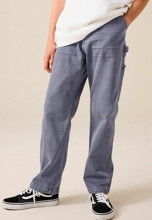 Personne portant un jean gris ample avec des passants latéraux et des baskets noires et blanches, debout avec une main dans la poche.