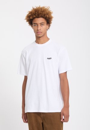 Jeune homme aux cheveux bouclés portant un T-shirt blanc uni avec un petit logo sur la poitrine et un pantalon en velours côtelé marron.
